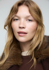 Giovane donna con capelli biondi chiari e lentiggini indossa un cappotto marrone con un colletto di pelliccia spesso, guardando con calma verso la fotocamera.