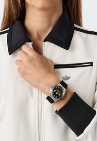 Hõbedane kell musta ketiga ja kuldsete aktsentidega, millel on kummist rihm. Kantud käel, millele sobib valge ja musta värvi lukuga jope.