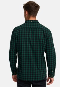 Zeleno-černá kostkovaná flanelová košile s dlouhými rukávy, zapínacími manžetami a límcem. Uvnitř zadní části je závěsný poutko.