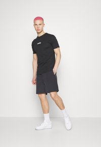 Black short-sleeve t-shirt with white logo, paired with dark shorts, white sneakers, and white socks. Model stands against a plain background.