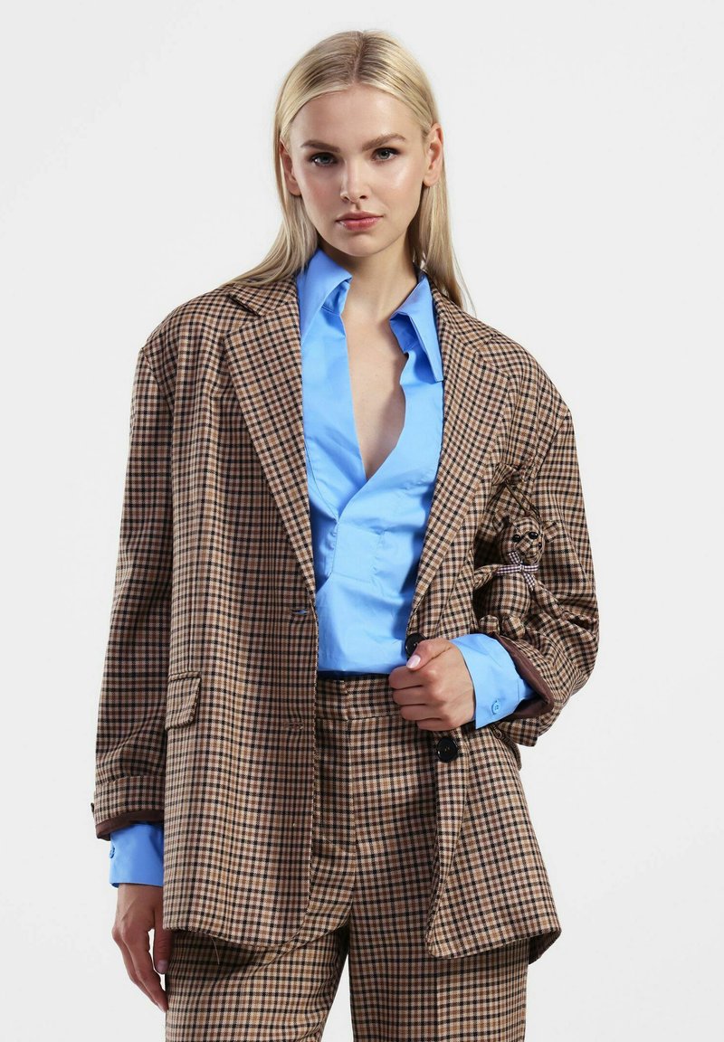 Blazer oversize a scacchi marroni sopra una camicia blu con bottoni, abbinato a pantaloni coordinati. Presenta una vestibilità rilassata e grandi bottoni.