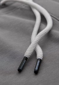 Grijs stof met een gladde textuur heeft twee witte katoenen trekkoorden, elk eindigend in zwarte plastic aglets gemerkt "DMBRE."