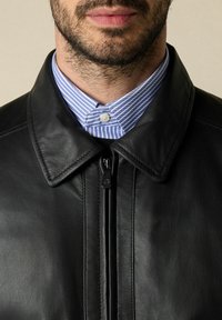 Man met een kort getrimde baard die een zwart leren jack draagt, gesloten boven een blauw-wit gestreept overhemd met de kraag dichtgeknoopt.