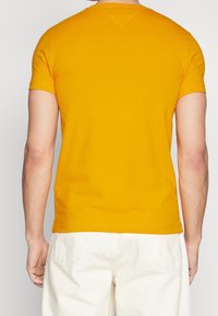 Man som bär en kortärmad senapsgul t-shirt och off-white byxor, sedd bakifrån mot en enfärgad grå bakgrund.