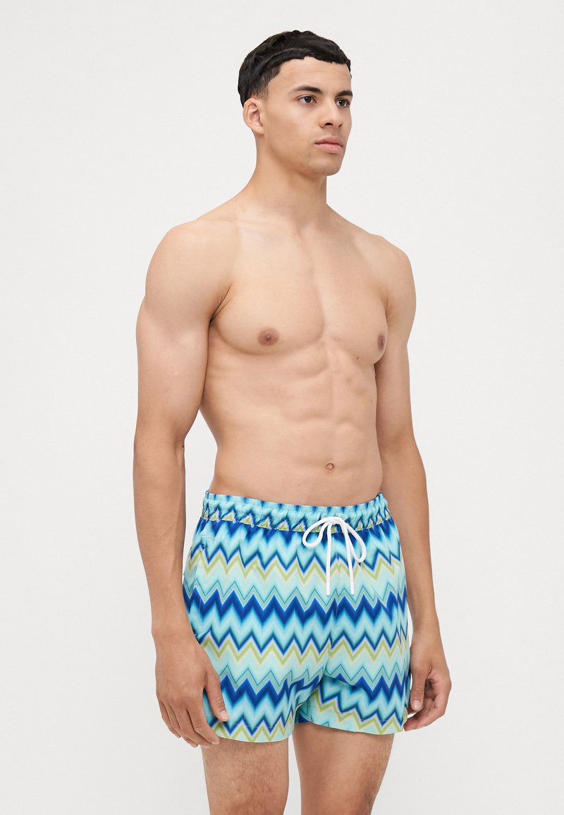 Costumi da bagno con un motivo a zigzag in tonalità di blu, verde e bianco. Presentano una vita elasticizzata con coulisse e una vestibilità comoda.