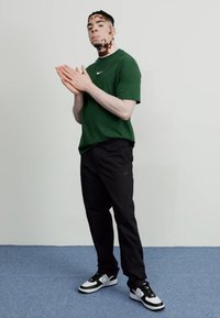 Grön kortärmad Nike-tröja, svarta Nike-byxor och svarta och vita sneakers. Modellen står på en blå matta med en neutral bakgrund.