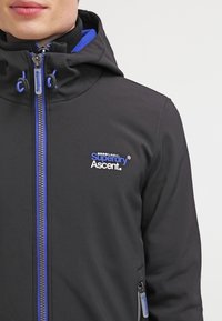 Svart jacka med en figursydd design, kontrasterande blå dragkedja och detaljer. Har en huva och broderad logotyp på bröstet.