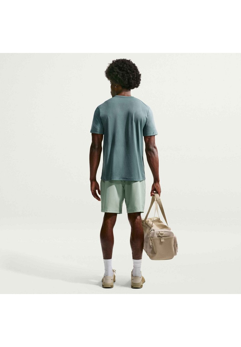 Homme vu de dos portant un t-shirt vert, un short vert clair, des chaussettes blanches, des baskets beiges, tenant un sac de sport beige dans la main droite.