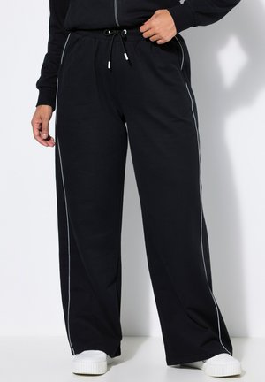 Persona con pantalones negros de pierna ancha con franjas blancas a los lados, una camiseta negra y zapatillas blancas, de pie frente a un fondo liso.