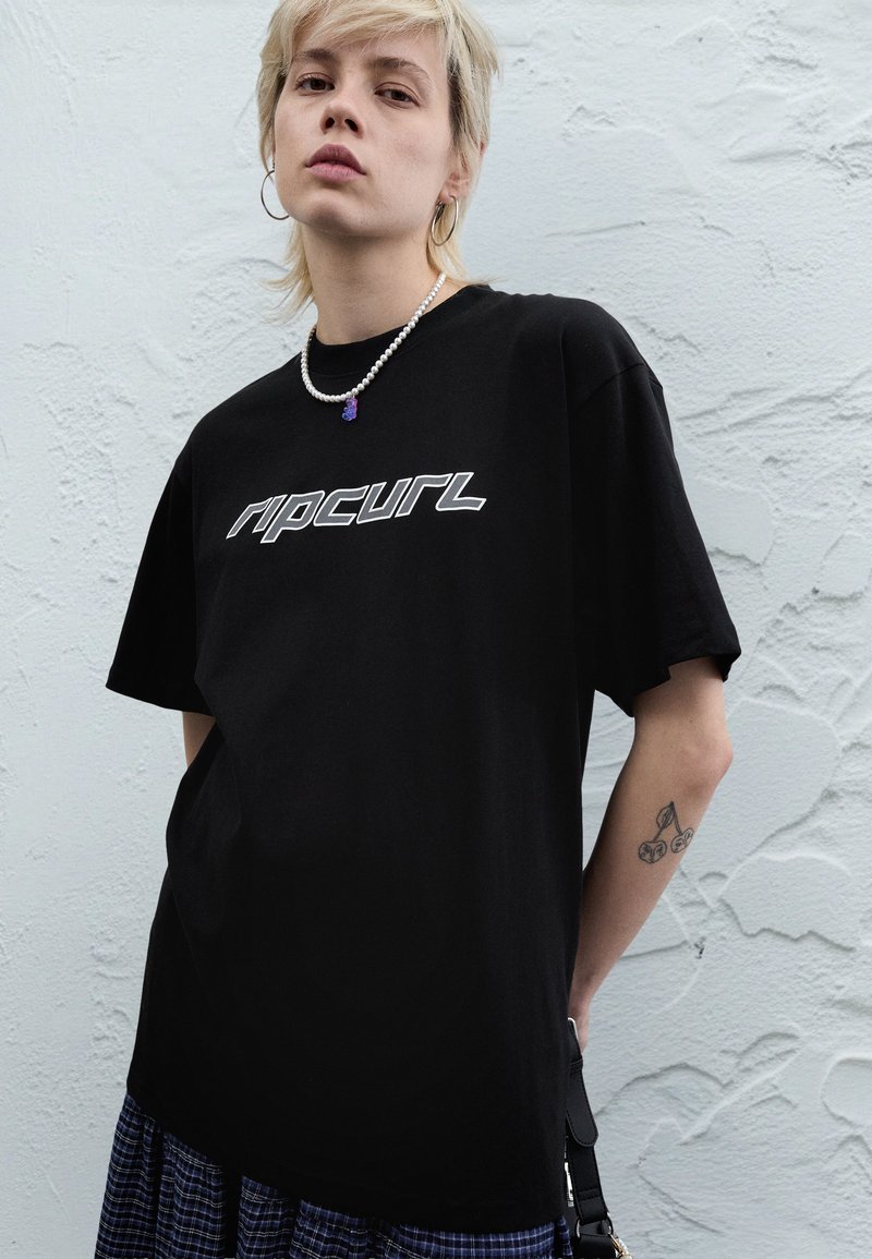 Black cotton t-shirt featuring white "ripcurl" logo. Short sleeves, relaxed fit. Model wears pearl necklace and displays arm tattoo.