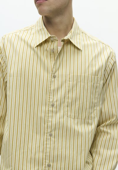 Vaaleankeltainen nappipaita, jossa ohuet pystysuorat raidat tummemmassa keltaisessa. Siinä on terävä kaulus ja rintatasku. Sileä pinta.