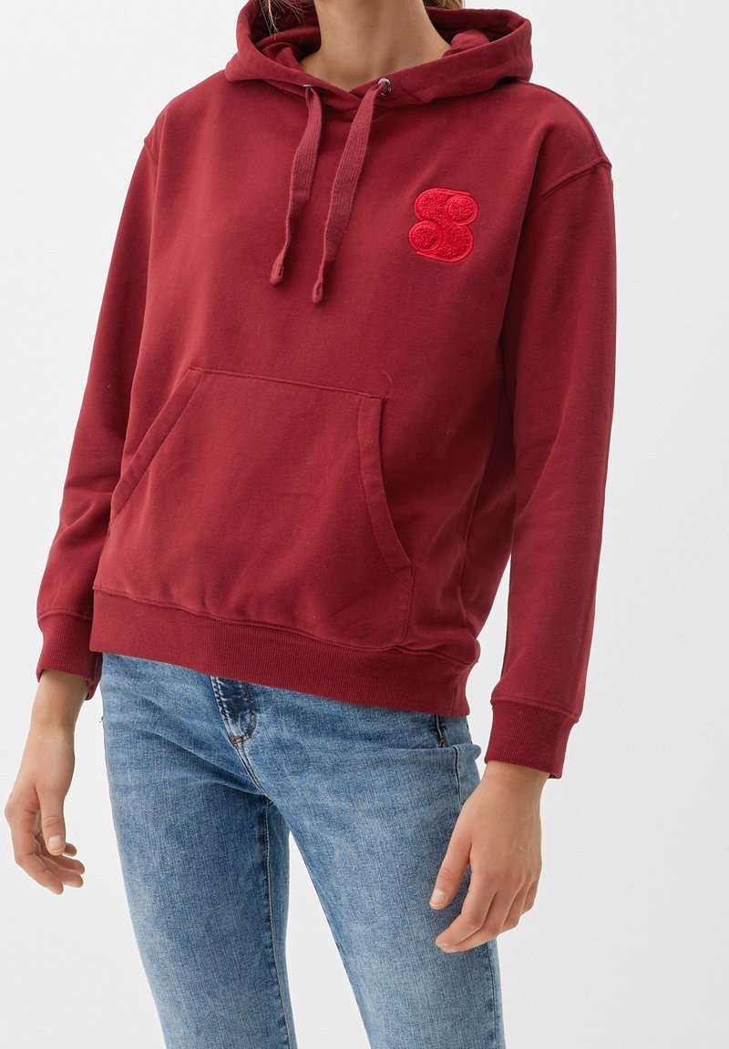 Sweat à capuche bordeaux avec poche avant et logo brodé rouge, doté de poignets et d'un ourlet côtelés, assorti à un jean bleu clair.