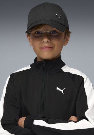 Schwarzer Puma Trainingsanzug mit weißen Akzenten, Reißverschluss vorne und Logo. Schwarze Mütze und transparente Brille runden den Look ab. Glatte Stoffoberfläche.