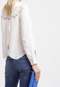 Chemisier blanc à manches longues avec des détails en dentelle sur les épaules, associé à un jean bleu foncé et une pochette bleue. Le chemisier a un dos fendu.