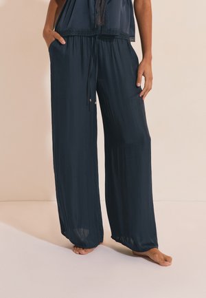 Kobieta ubrana w luźne, ciemnogranatowe spodnie o szerokich nogawkach z kieszeniami i wiązanym w pasie sznurkiem, stojąca boso na neutralnym tle.