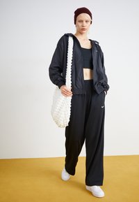Mustat kevyet takit, crop top ja löysät housut yhdistettyinä valkoiseen paksutekstuuriseen laukkuun ja valkoisiin lenkkareihin keltaisella lattialla.