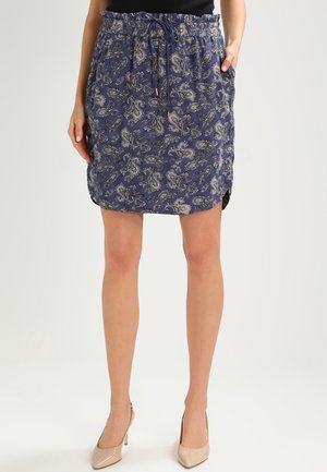 Vrouw met een blauwe rok met paisley-patroon, zakken en een taille met koord, gecombineerd met beige pumps.