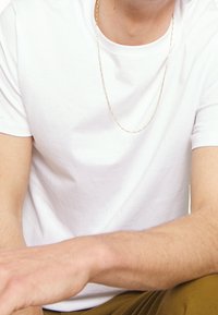 T-shirt en coton blanc avec un col rond, texture lisse, porté avec un collier en chaîne dorée fin, mains et bras partiellement visibles.