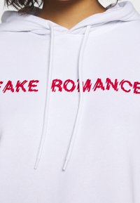 Vit hoodie med "FAKE ROMANCE" i fet, röd, sliten text över framsidan. Har snören och är tillverkad av mjukt material.