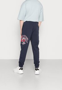 Tmavě modré tepláky s výrazným červeným logem "TOMMY" na levé straně, doplněné o pohodlný střih a manžety na zápěstí. Kombinované s bílými teniskami.