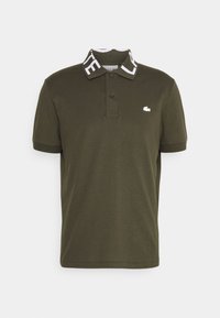 Olivově zelené polo tričko vyrobené z bavlny, s kontrastním bílým límcem s nápisem "LACOSTE" a malou bílou logem na hrudi.