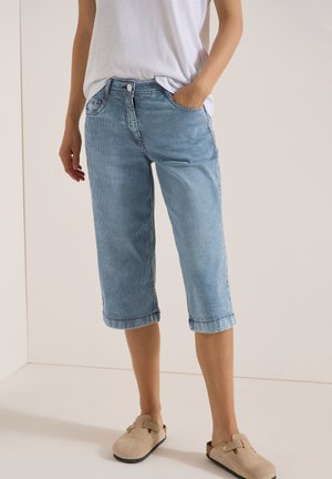 Persoon die een wit T-shirt, blauw-wit gestreepte knielange spijkershort en beige instapschoenen draagt, staat met één hand in de zak.
