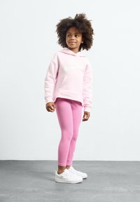 Roze dukserica s bijelim Nike logom, uparen s roza tajicama i bijelim tenisicama. Glatka tekstura i ležeran kroj. Nema dodatnih detalja.