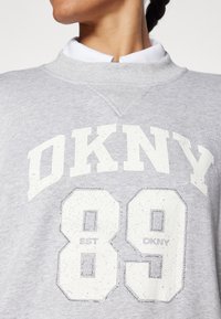 Pilkas džemperis su apvalia kaklo iškirpte, su „DKNY“ ir „89“ baltos spalvos su blizgiu akcentu. Medvilnės mišinys su lygiu tekstūru.