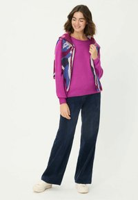 Lila gestrickter Pullover mit bunter, gemusterter Weste darüber. Kombiniert mit dunkelblauen Cordhosen und weißen Turnschuhen.