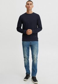 Pulover bleumarin cu mâneci lungi, guler rotund și mic detaliu cu logo la tiv, purtat cu blugi albaștri deschiși și adidași negri.