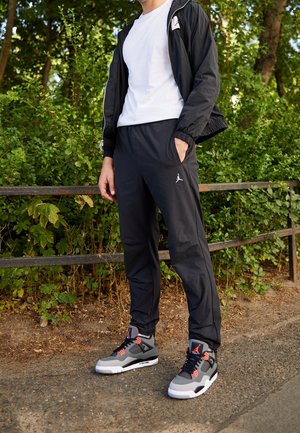 Mand iført sorte træningsbukser, sort jakke, hvid t-shirt og grå, sorte og orange sneakers, stående på en sti ved grønne buske.