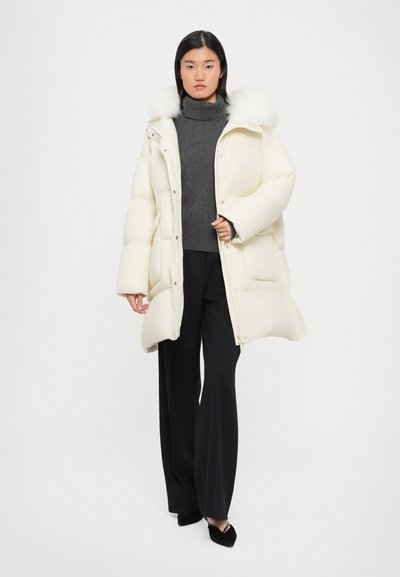 Weißer Puffer-Mantel mit einer Kapuze aus Kunstfell, Reißverschluss vorne und großen Taschen. Kombiniert mit einem grauen Rollkragenpullover und schwarzen weit geschnittenen Hosen. Glatte Textur.