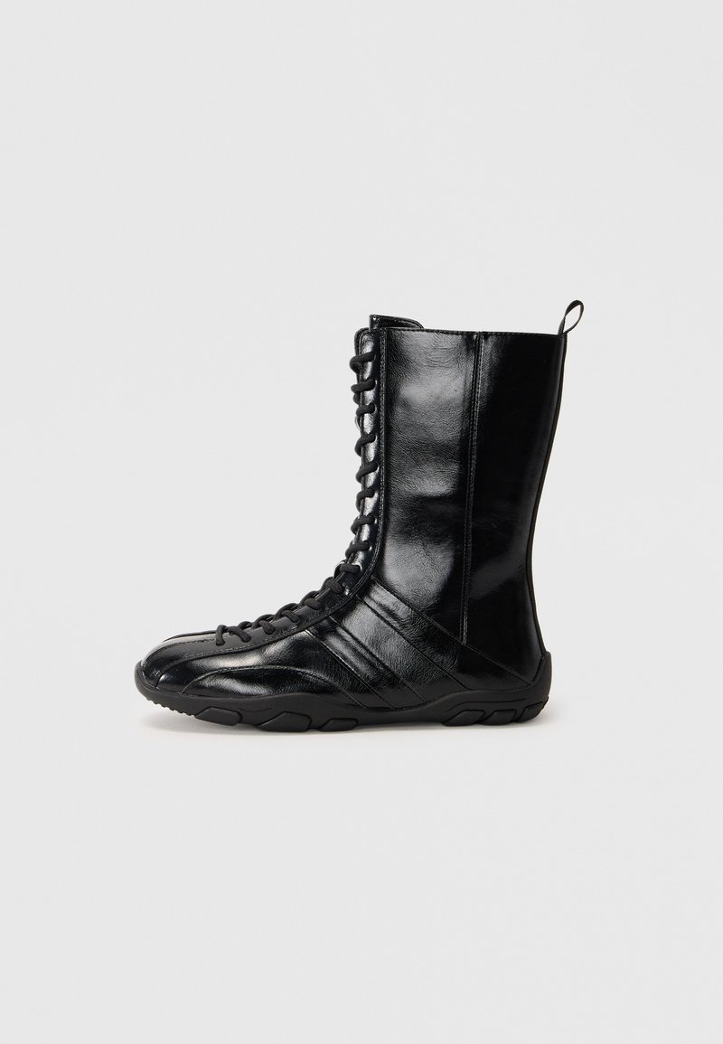 Botas de cuero negras con caña alta, cordones en la parte delantera, suela de goma y paneles texturizados. Cuenta con un lazo para facilitar su uso.