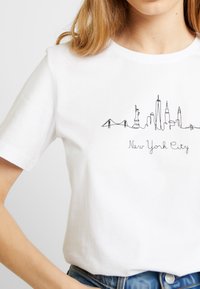 Bílá bavlněná tričko s černým liniovým uměním skyline New Yorku, s textem pod ním, který zní "New York City". Jednoduchý kulatý výstřih.