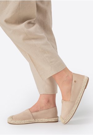 Beige Espadrilles zum Hineinschlüpfen mit gewebter Jutesohle, getragen mit hellbeigen, verkürzten Leinenhosen vor weißem Hintergrund.