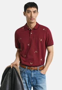 Mann in burgunderfarbenem Polo mit Vogelmotiven, blauen Jeans, geflochtenem braunen Gürtel, hält eine schwarze Lammfelljacke in einer Hand, die andere Hand in der Tasche.