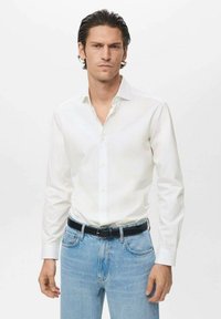 Mand iført en hvid skjorte med knapper, stoppet ned i lyseblå jeans med et sort bælte, stående op ad en ensfarvet hvid baggrund.
