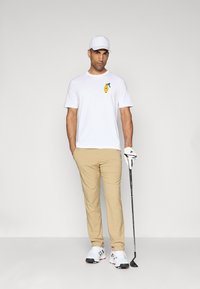 White t-shirt with green and yellow logo, khaki pants, white baseball cap, and golf shoes. Holding a golf club, standing on neutral background.