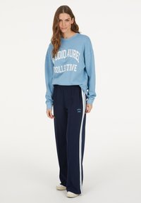 Blauer Baumwollpullover mit weißem Text, kombiniert mit marineblauen Hosen mit weißen Seitestreifen, getragen mit weißen Turnschuhen.