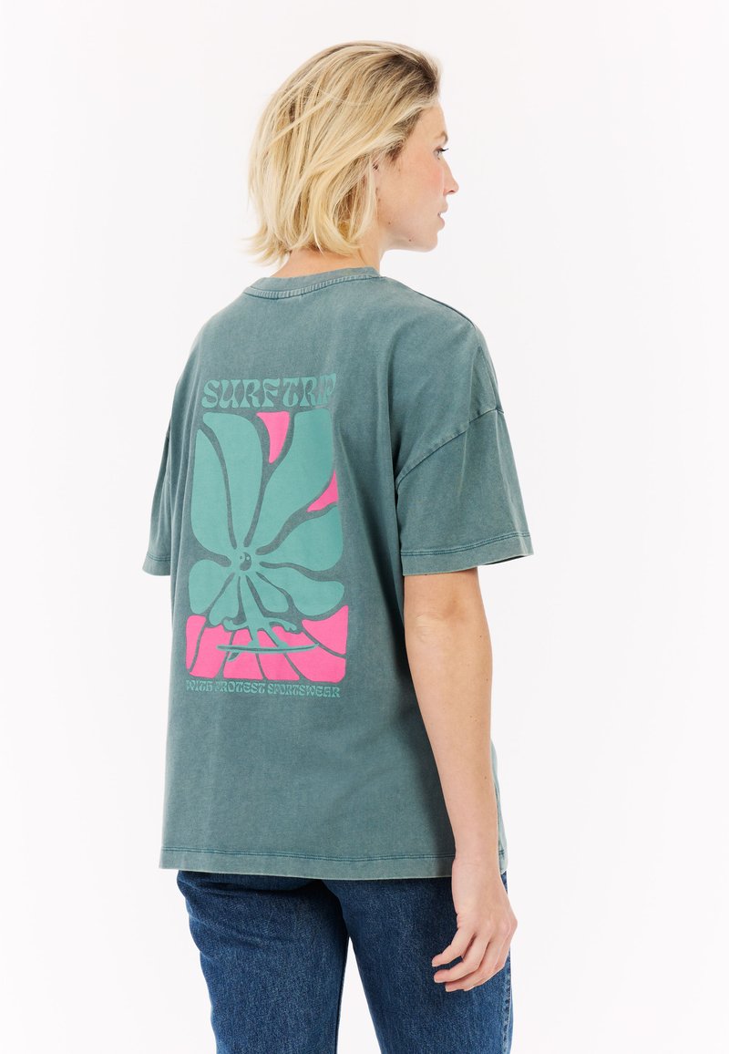 Overgrote groene katoenen t-shirt met een grote roze en teal bloemenafbeelding op de achterkant, met daarboven de tekst "SURFTRIP". Klassieke ronde hals.