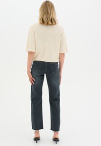 Suéter de punto beige con mangas cortas, combinado con jeans de mezclilla oscuros con dobladillo deshilachado. El modelo es visto por la espalda.