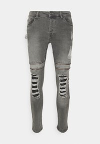 Jeans de denim gris con un ajuste slim, detalles desgastados y rasgaduras a rayas negras a lo largo de las piernas. Acentos de cremallera y bordes deshilachados.