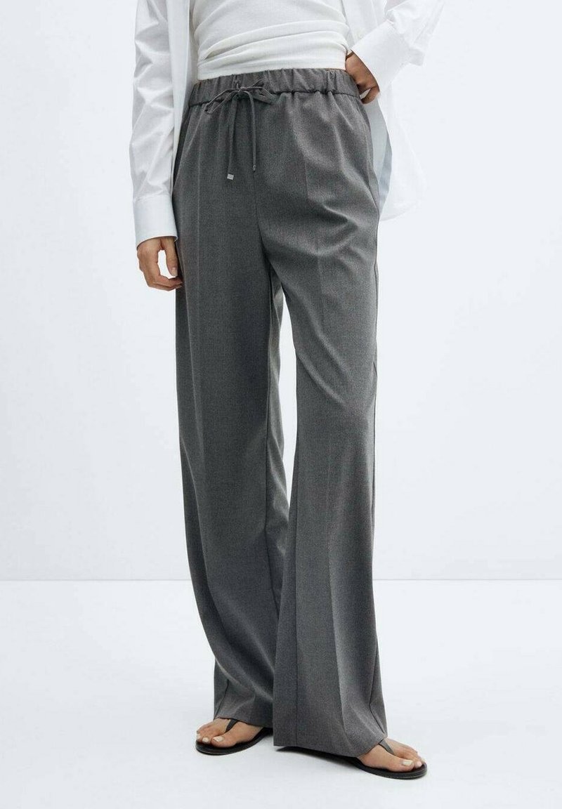 Personne portant un pantalon gris foncé à jambes larges avec une taille à cordon, associé à une chemise blanche et des sandales noires avec des brides au niveau des orteils.