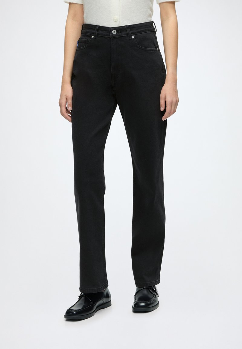 Person iført lige sorte jeans parret med sorte pæne sko og en cremefarvet kortærmet knappet top, stående mod en hvid baggrund.