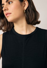 Zwarte mouwloze top met een ronde halslijn en zichtbare naaddetails, gemaakt van een gladde, zachte stof. Gouden hoepeloorbel zichtbaar.