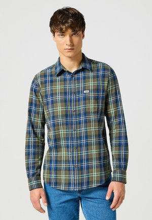Giovane uomo che indossa una camicia a scacchi verde e blu con taschino e jeans blu, in piedi contro uno sfondo chiaro e uniforme.