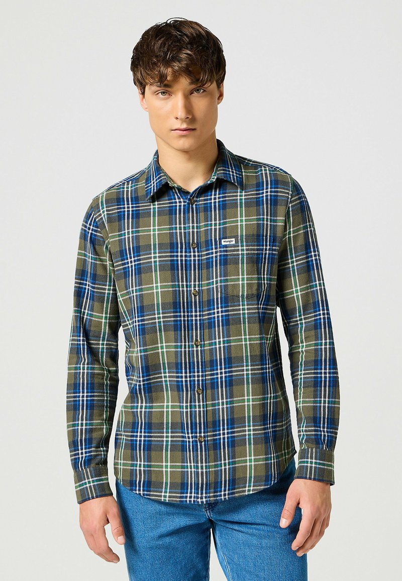 Giovane uomo che indossa una camicia a scacchi verde e blu con taschino e jeans blu, in piedi contro uno sfondo chiaro e uniforme.