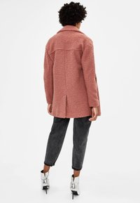 Cappotto rosa strutturato con colletto, tasche laterali e taglio dritto, abbinato a jeans scuri con risvolto e stivaletti argentati lucidi.