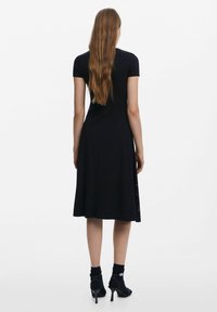 Robe noire à manches courtes avec une jupe évasée, fabriquée en matériau doux ; possède un imprimé décoratif le long de l'ourlet, associée à des chaussures à talons hauts noires.