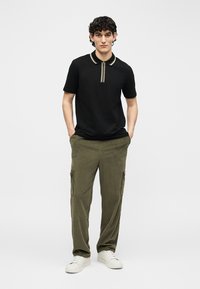 Jeune homme portant un polo noir zippé, un pantalon cargo en velours côtelé vert olive et des baskets blanches, debout les mains dans les poches.